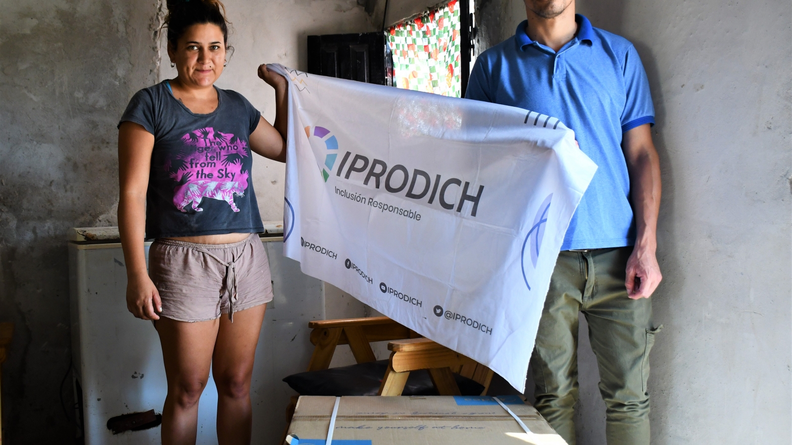 Personal del IPRODICH sosteniendo bandera institucional frente a cámara junto a beneficiaria, que tiene a sus pies un aire acondicionado recibido